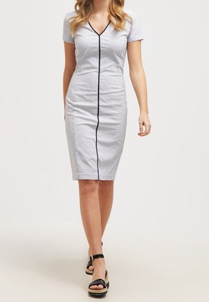 Kvinde gående iført en tætsiddende, kortærmet, knælængde lysegrå kjole med en sort lodret stribe ned i midten og sorte platåsandaler.