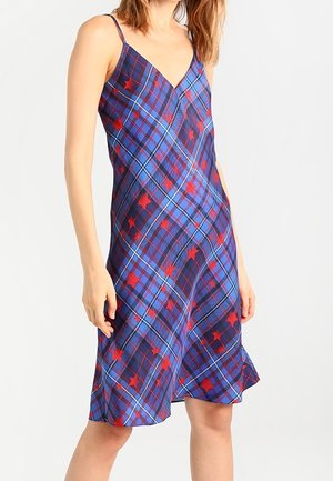 Femme portant une robe sans manches à col en V, à carreaux bleus avec des motifs d'étoiles rouges, debout devant un fond blanc uni.