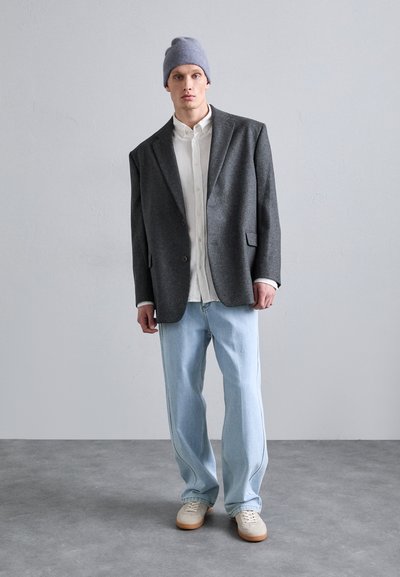 Blazer grigio oversize, camicia bianca con bottoni, jeans azzurri chiari, sneakers crema e un berretto grigio chiaro. Tessuti morbidi e vestibilità rilassata.