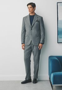 Grau gemusterter Anzug mit tailliertem Sakko und Hose, ergänzt durch einen dunkelblauen Pullover und schwarze Schuhe. Verfügt über ein Einstecktuch.