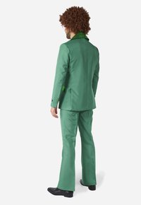 Costume sur mesure vert avec un col texturé, pantalons évasés et matériau lisse. Les accessoires incluent des chaussures noires brillantes et une perruque brune bouclée.