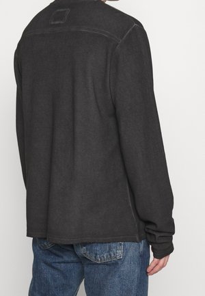 Uomo che indossa una camicia scura a maniche lunghe e jeans blu, mostrato di spalle contro uno sfondo chiaro e semplice.