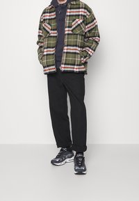 Zielona koszula flanelowa w kratę z czerwonymi i białymi akcentami, nałożona na ciemny hoodie. Czarne spodnie i czarne buty sportowe dopełniają strój.