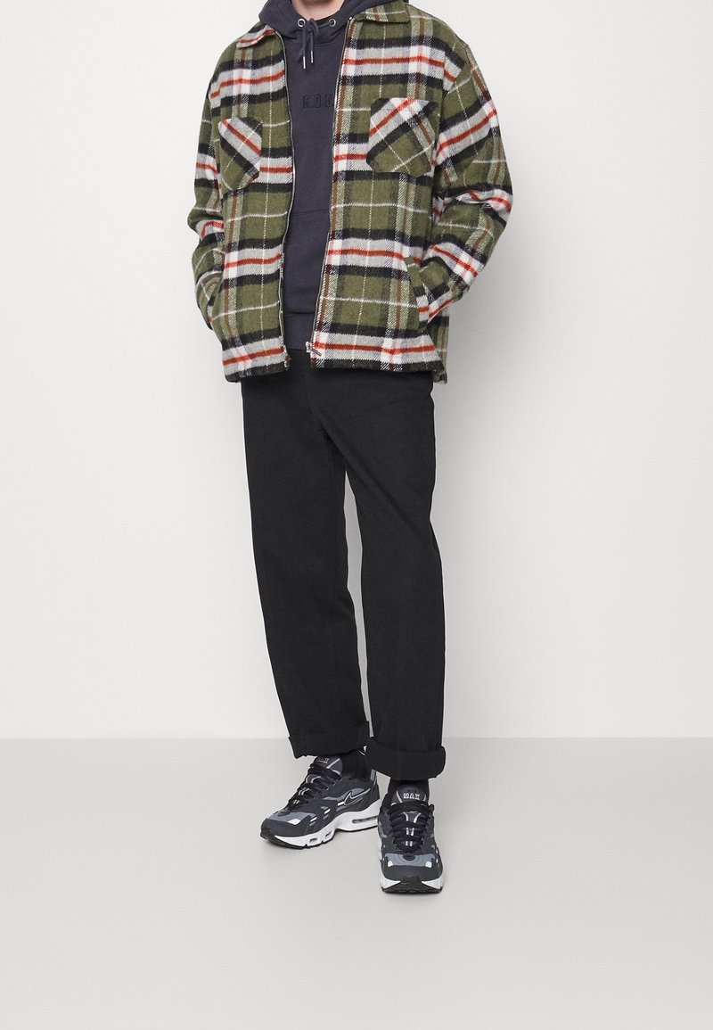 Zielona koszula flanelowa w kratę z czerwonymi i białymi akcentami, nałożona na ciemny hoodie. Czarne spodnie i czarne buty sportowe dopełniają strój.