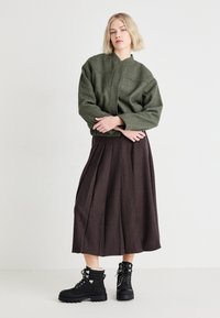 Veste en laine texturée vert olive avec col montant, associée à une longue jupe plissée marron et des bottes noires à lacets avec doublure en fourrure.