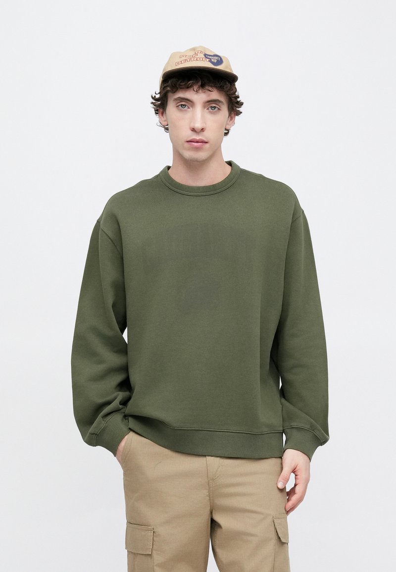Green sweatshirt with a crew neck and long sleeves. The fabric appears soft and textured. Paired with beige cargo pants.