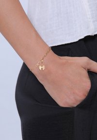 Bracelet en or avec un charme plat en forme de nœud sur une chaîne. La main repose sur une poche de pantalons noirs, avec un haut blanc visible.