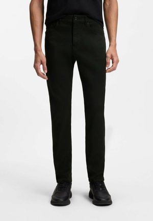 Mann trägt schlanke schwarze Hose, schwarze Sneakers und schwarzes T-Shirt, steht vor weißem Hintergrund.