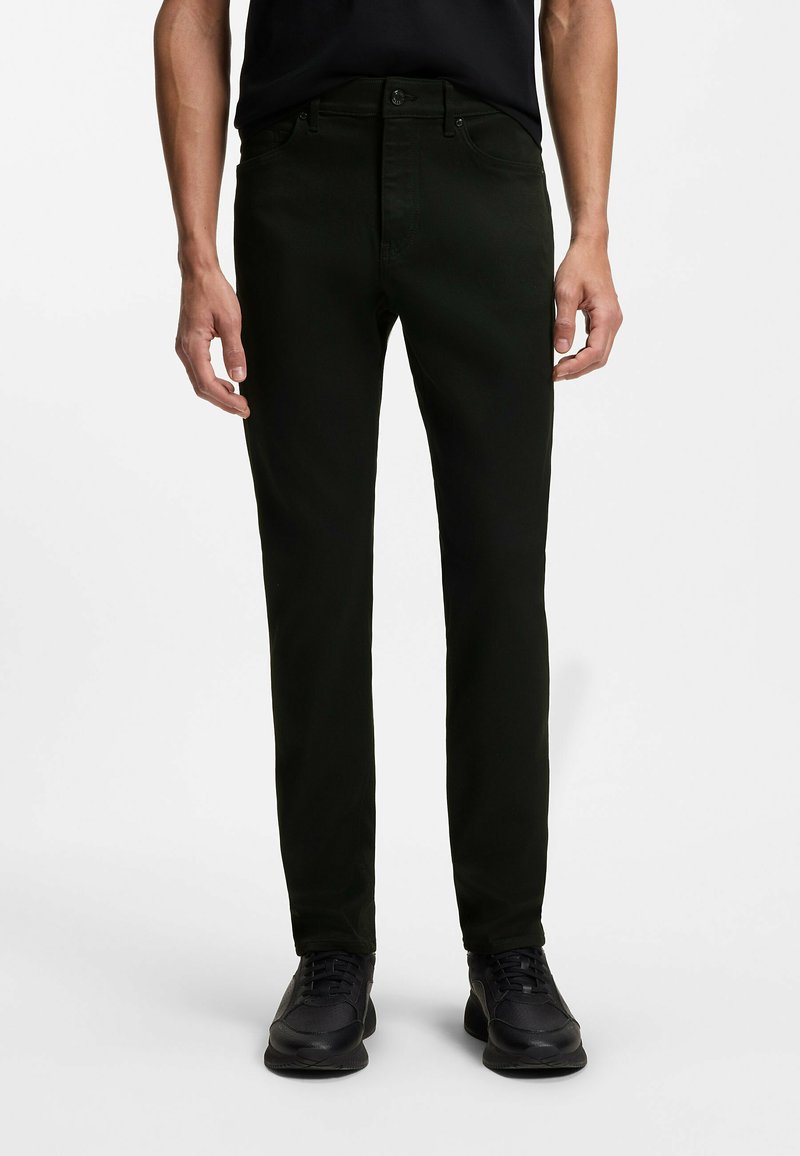 Mężczyzna ubrany w dopasowane czarne spodnie, czarne tenisówki i czarną koszulkę, stojący na białym tle.