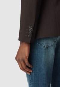 Brązowa kurtka w kratę z klapą, z czterema czarnymi guzikami na mankietach, noszona z niebieskimi dżinsami. Widoczna teksturowana tkanina.