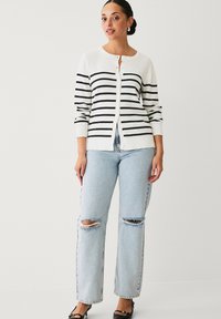 Cardigan bianco con strisce orizzontali nere, dotato di maniche lunghe e chiusura con bottoni. Abbinato a jeans strappati blu chiaro e scarpe nere.