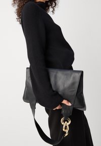 Sac à bandoulière en cuir noir de forme triangulaire, avec des ferrures dorées et une bandoulière amovible, tenu par une personne portant un pull noir à côtes.
