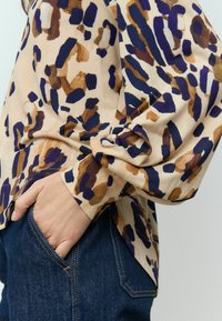 Blouse beige avec des motifs abstraits marron et marine, à manches longues et à coupe décontractée, associée à un jean bleu foncé.