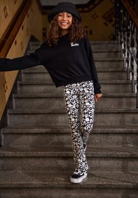 Fekete cropped pulóver fehér logóval, fekete-fehér mintás harangszárú nadrággal és fekete sportcipővel, kő lépcsőn.