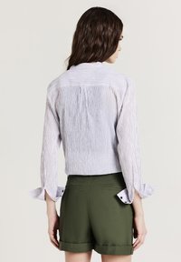 Camicia a righe azzurro chiaro e bianca con schiena plissettata, maniche lunghe e polsini arrotolati, abbinata a pantaloni corti verde oliva con tasche.