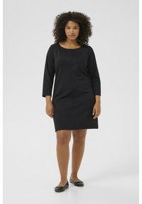 Robe noire à manches longues avec un col rond, détails de plis à l'avant et coupe droite. Portée avec des ballerines noires, design simple.