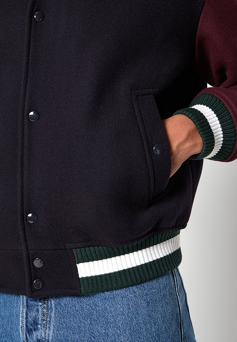 Veste de varsity noire et bordeaux avec boutons-pression, poignets côtelés verts et blancs, dotée d'une poche latérale et d'un tissu texturé.