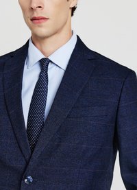 Blazer blu navy con trama, revers a lancia, chiusura con un bottone, indossato sopra una camicia da uomo a righe azzurre e una cravatta fantasia.