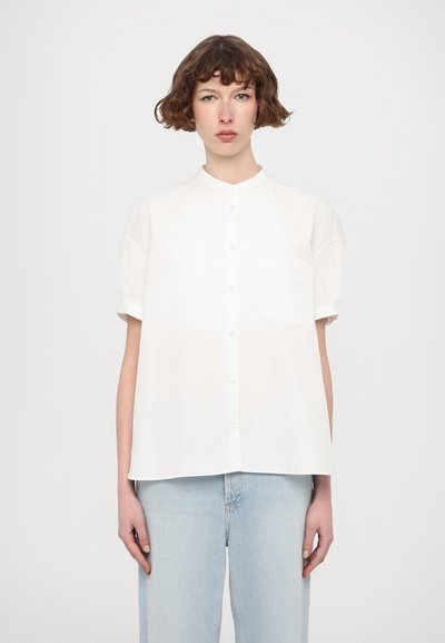 Chemise blanche à manches courtes avec un col rond, des boutons sur le devant et une poche poitrine. Tissu léger, texture lisse, portée avec un jean ample bleu clair.