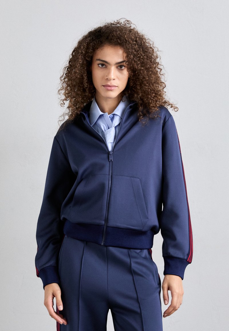 Giacca navy con zip, polsini e orlo a coste, presenta strisce laterali rosse, tasche frontali e una texture di tessuto liscia e leggermente lucida.
