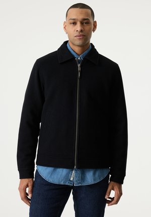 Man standing facing forward, wearing a black zip-up jacket over a blue button-up shirt and dark jeans against a plain background.