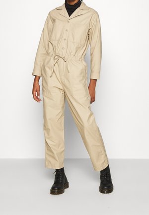 Combinaison beige à manches longues, devant avec boutons, deux poches poitrine, taille élastiquée et jambes fuselées. Portée avec des bottes noires à lacets.