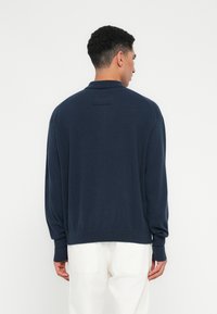 Uomo di spalle che indossa un maglione lungo blu scuro e pantaloni bianchi su uno sfondo chiaro semplice.