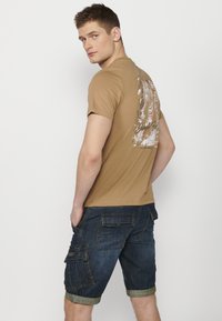 T-shirt en coton marron avec un imprimé graphique au dos, associé à un short cargo en denim bleu foncé avec de multiples poches et des revers au niveau des cuisses.