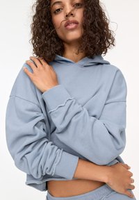 Sweat-shirt court bleu clair avec capuche, coupe ample et poignets côtelés. Tissu doux avec des détails de couture visibles à l'ourlet et aux manches.