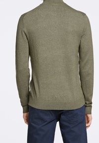 Pull en maille verte avec un col montant, des poignets et un ourlet côtelés. Texture lisse, design ajusté, associé à un pantalon bleu foncé.