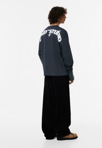 Camicia a maniche lunghe di colore grigio scuro con grande testo bianco sul retro, abbinata a pantaloni in velluto a coste neri a gamba larga e calzature casual.