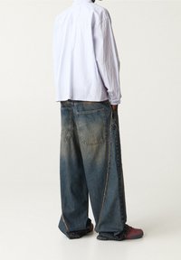 Luźne niebieskie dżinsy o wyblakłej fakturze i kontrastowych szwach, zestawione z jasnofioletową koszulą z długim rękawem i wielokolorowym obuwiem.