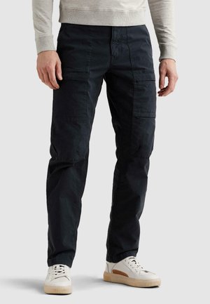 Mężczyzna w czarnych spodniach typu cargo i białych trampkach, stojący na tle jasnego, jednolitego tła, z rękami swobodnie opuszczonymi wzdłuż ciała.