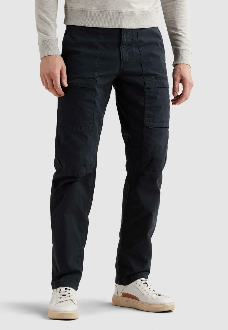Homme portant un pantalon cargo noir et des baskets blanches, debout contre un fond clair uni les mains détendues le long du corps.