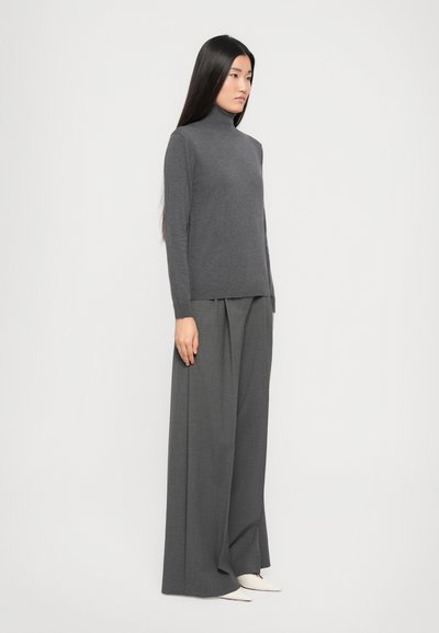 Femme avec de longs cheveux noirs portant un pull à col roulé gris, un pantalon gris à jambes larges et des chaussures blanches à bout pointu sur un fond blanc.