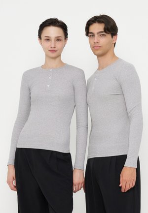 Zwei Personen stehen nebeneinander und tragen hellgraue langärmelige Henley-Shirts und schwarze Hosen vor einem einfarbigen weißen Hintergrund.