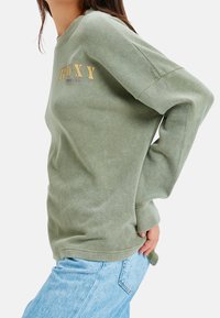 Personne portant un pull en maille à manches longues vert clair avec le texte "ROXY" et un jean bleu clair, posant avec une main derrière le dos.
