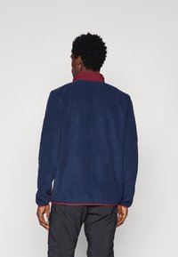 Giacca in pile blu navy con colletto bordeaux, caratterizzata da una vestibilità comoda e una texture liscia, cuciture visibili all'orlo e maniche lunghe.