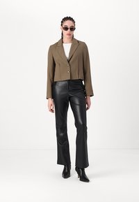 Blazer vert olive coupé avec un bouton unique, associé à un pantalon évasé en simili cuir noir et des bottines à talons noires.