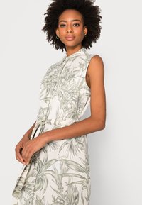 Abito senza maniche in tessuto leggero con motivo a foglie tropicali verdi, chiusura frontale con bottoni, vita accentuata da una cintura e dettagli con spacco laterale.