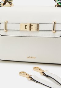 Valkoinen nahkainen käsilaukku, jossa kullanväriset metalliosat, teksturoitu pinta, lukollinen suljin ja irrotettavat hihnat. Brändinimi "ALDO" näkyvissä.