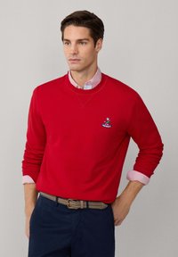 Felpa rossa in cotone con polsini a costine, caratterizzata da un piccolo logo. Indossata sopra una camicia colletto a righe rosa chiaro e bianca. Pantaloni blu navy.