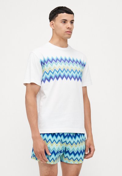 T-shirt blanche avec un motif en zigzag bleu et vert sur la poitrine, associée à un short à motifs multicolores. Manches courtes, coupe décontractée.