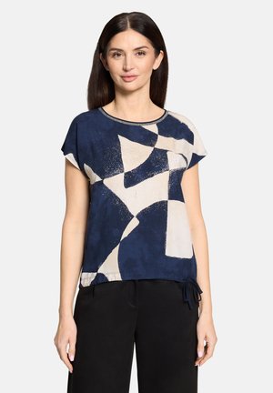 Kobieta ubrana w krótkim rękawem, granatowo-beżową bluzkę z abstrakcyjnym wzorem i okrągłym dekoltem oraz czarne spodnie, stojąca na jednolitym tle.