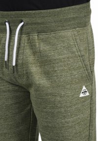 Grüne melierte Sweatpants mit einem gerippten Bund, weißen Kordeln und einem kleinen dreieckigen Logo im Bereich der linken Tasche.