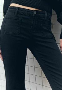 Un jean en denim noir présente une coupe slim, un design à cinq poches et des coutures visibles. Le tissu semble lisse avec une légère élasticité.