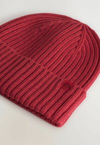 Bonnet en tricot côtelé rouge avec revers replié et petite étiquette en tissu arborant un logo de montagne sur fond uni.