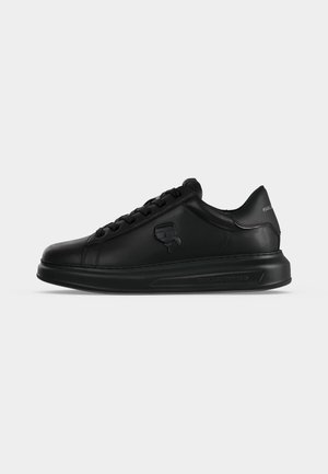 Zapatilla negra de piel de corte bajo con logo en relieve en el lateral y suela gruesa sobre un fondo gris liso.