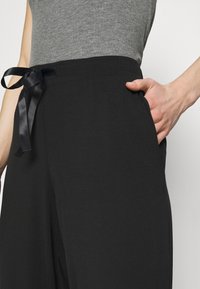 Zwarte wijde-pijp broeken met een gladde textuur, beschikt over een zijzak en een satijnen ceintuur op de taille, gedragen met een grijze nauwsluitende top.