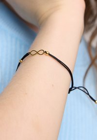 Czarny sznurkowy bransoletka z złotym symbolem nieskończoności i złotymi koralikami. Bransoletka jest regulowana i noszona na jasnej skórze.